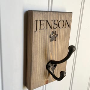 Pu&ograve; includere: Un gancio in legno per guinzaglio per cani con il nome "Jenson" inciso su di esso e un'impronta di zampa sotto il nome. Il gancio &egrave; fissato a una parete bianca.