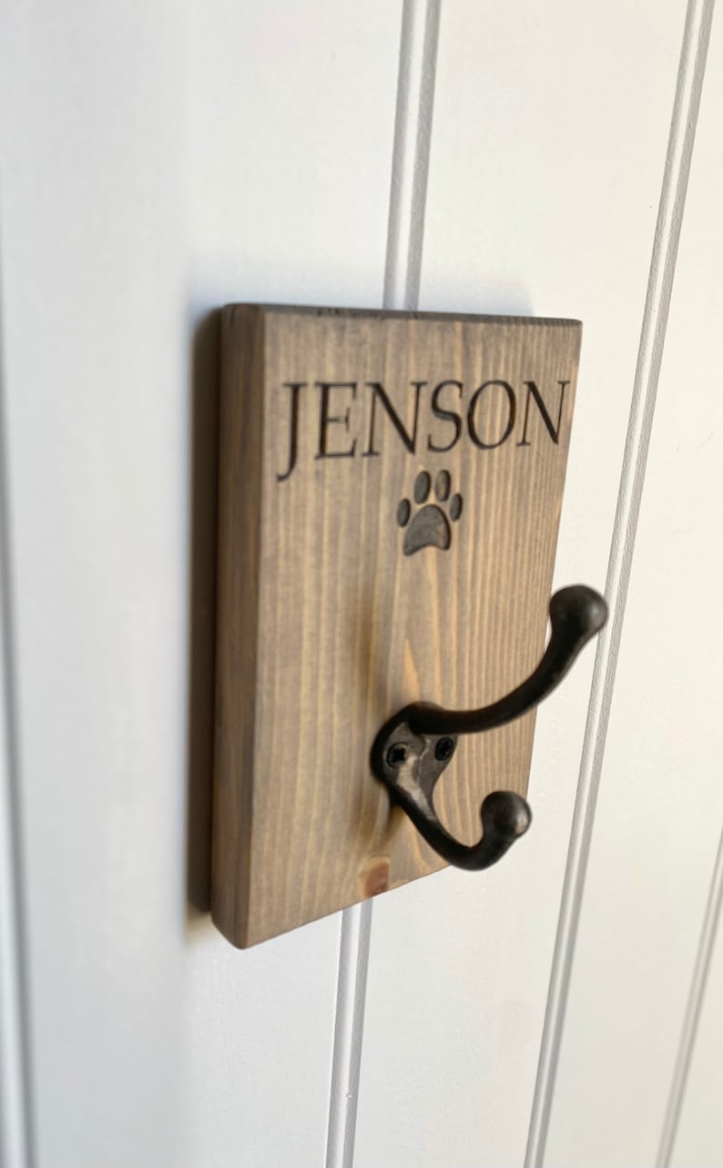 Pu&ograve; includere: Un gancio in legno per guinzaglio per cani con il nome "Jenson" inciso su di esso e un'impronta di zampa sotto il nome. Il gancio &egrave; fissato a una parete bianca.