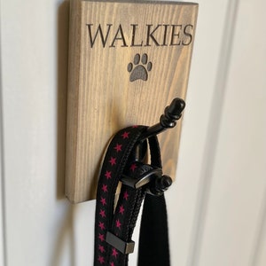 Pu&ograve; includere: Un cartello di legno con la scritta "Walkies" e un'impronta di zampa, con un gancio nero per appendere i guinzagli. Un guinzaglio nero con stelle rosa e un collare nero sono appesi al gancio.