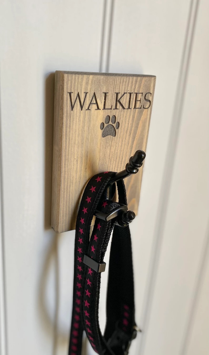 Pu&ograve; includere: Un cartello di legno con la scritta "Walkies" e un'impronta di zampa, con un gancio nero per appendere i guinzagli. Un guinzaglio nero con stelle rosa e un collare nero sono appesi al gancio.