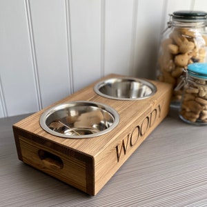 May include: A wooden pet food bowl stand with two stainless steel bowls. The stand is stained a dark brown and has the name "Woody" engraved on the front.