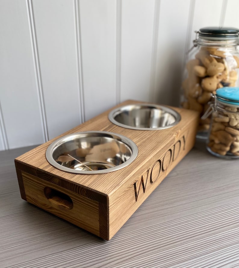 May include: A wooden pet food bowl stand with two stainless steel bowls. The stand is stained a dark brown and has the name "Woody" engraved on the front.
