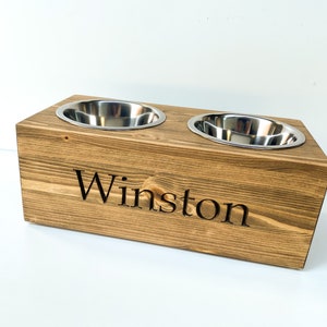 May include: A wooden elevated dog bowl stand with two stainless steel bowls. The stand is stained a dark brown and has the name "Winston" engraved on the front.