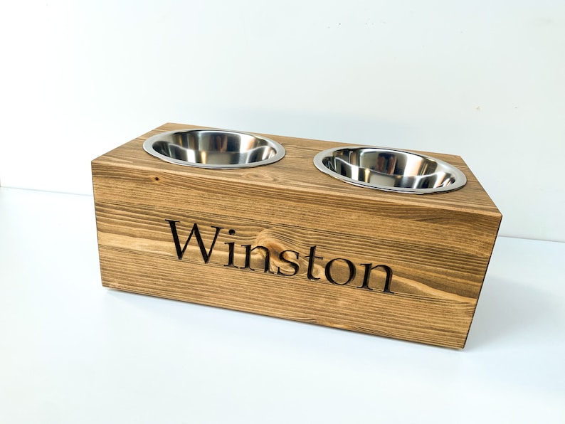 May include: A wooden elevated dog bowl stand with two stainless steel bowls. The stand is stained a dark brown and has the name "Winston" engraved on the front.