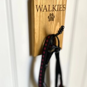 Pu&ograve; includere: Un gancio da parete in legno con la scritta "WALKIES" e un'impronta di zampa incisa su di esso. Un guinzaglio per cani nero e rosa &egrave; appeso al gancio.