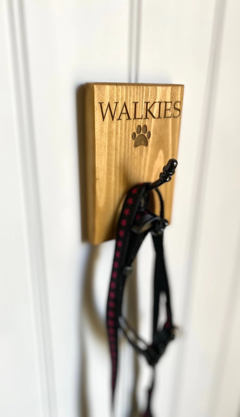 Pu&ograve; includere: Un gancio da parete in legno con la scritta "WALKIES" e un'impronta di zampa incisa su di esso. Un guinzaglio per cani nero e rosa &egrave; appeso al gancio.
