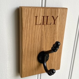 May include: A wooden wall hook with a black metal hook. The wood is light brown and has the name "LILY" engraved on it.