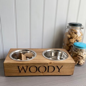May include: A wooden double dog bowl stand with two stainless steel bowls. The stand is stained a dark brown and has the name "WOODY" engraved on the front.