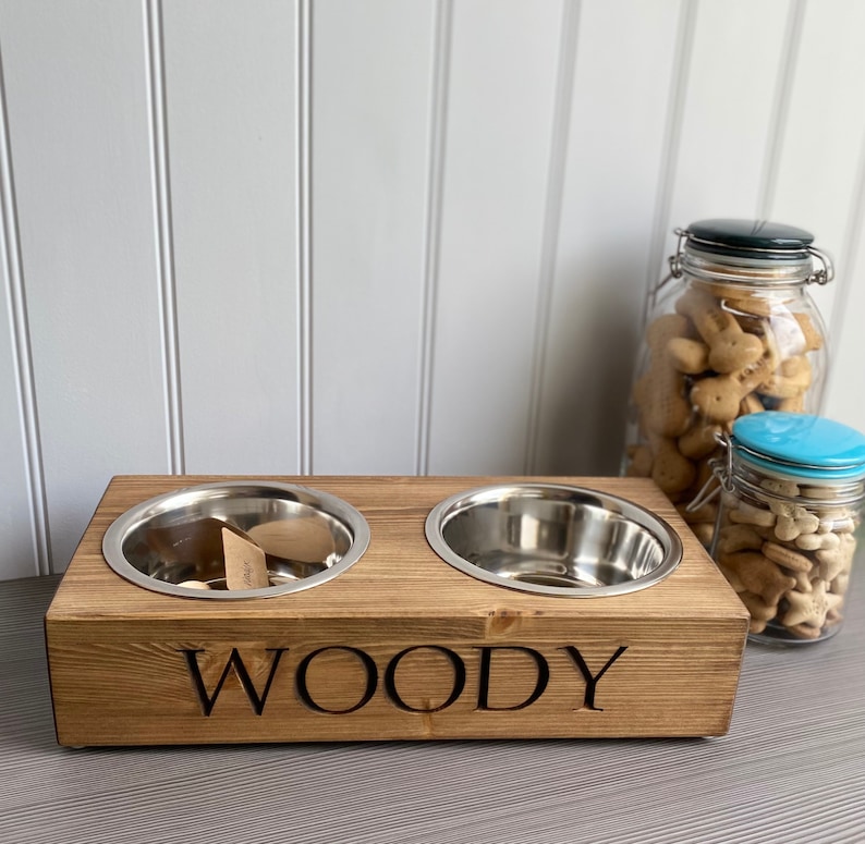 May include: A wooden double dog bowl stand with two stainless steel bowls. The stand is stained a dark brown and has the name "WOODY" engraved on the front.