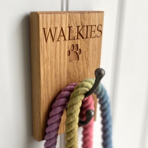 May include: A wooden wall-mounted hook with the word "WALKIES" engraved on it. The hook has a paw print design and is holding three colourful rope leashes.