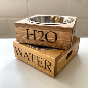 May include: Two wooden pet bowls with stainless steel inserts. The top bowl has "H2O" engraved on the front and the bottom bowl has "WATER" engraved on the front.