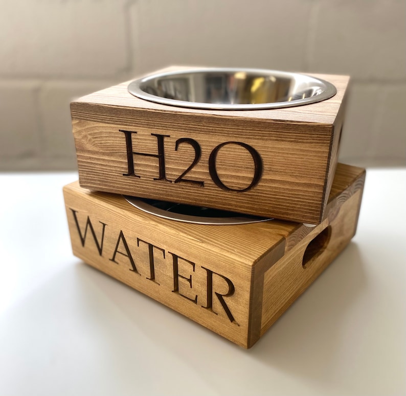 May include: Two wooden pet bowls with stainless steel inserts. The top bowl has "H2O" engraved on the front and the bottom bowl has "WATER" engraved on the front.