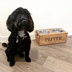 May include: A black dog sits in front of a wooden dog bowl stand with two stainless steel bowls. The stand is engraved with the name "PEPPER".