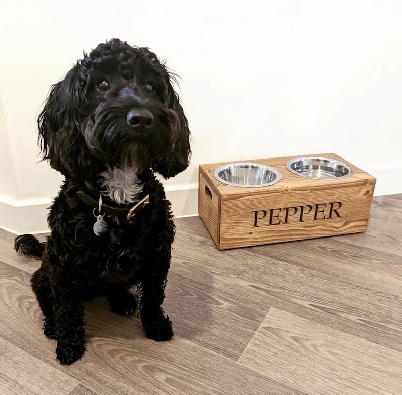 May include: A black dog sits in front of a wooden dog bowl stand with two stainless steel bowls. The stand is engraved with the name "PEPPER".