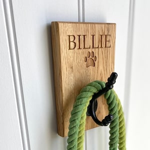 May include: A wooden dog leash hook with the name "Billie" engraved on it. A green leash is hanging from the hook. The hook is mounted on a white wall.