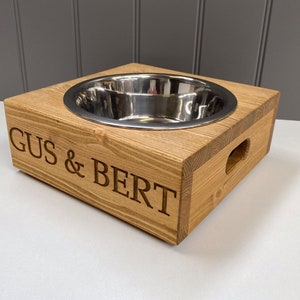May include: A wooden dog bowl stand with a stainless steel bowl. The stand is engraved with the names "Gus & Bert".