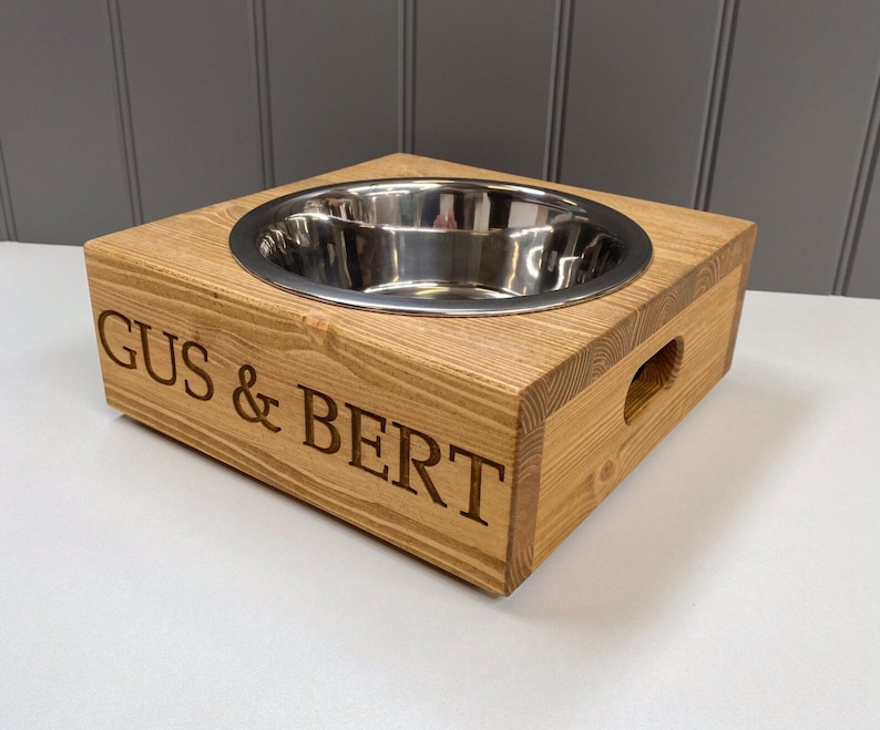 May include: A wooden dog bowl stand with a stainless steel bowl. The stand is engraved with the names "Gus & Bert".