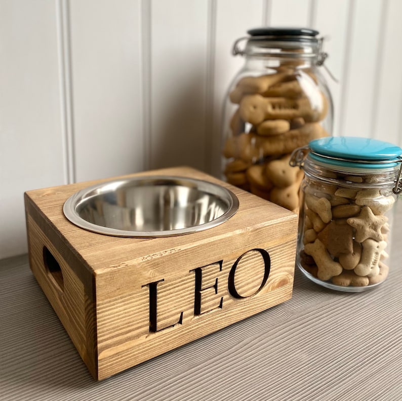 May include: A wooden dog bowl stand with a stainless steel bowl and the name "LEO" engraved on the front. The stand is made of natural wood and has a rustic look. Two glass jars filled with dog treats are in the background.