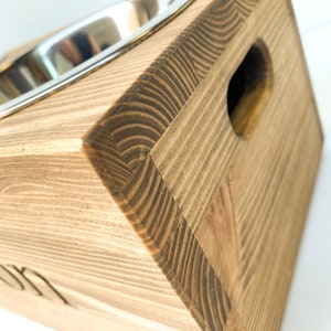 May include: A wooden pet food stand with two stainless steel bowls. The stand is stained a dark brown color and has a cutout on the side. The stand is engraved with the word "Winston".