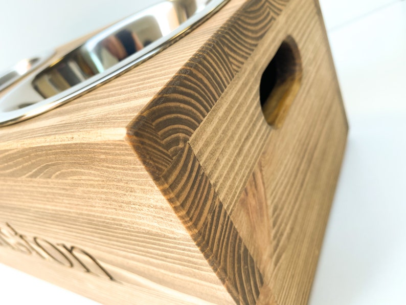 May include: A wooden pet food stand with two stainless steel bowls. The stand is stained a dark brown color and has a cutout on the side. The stand is engraved with the word "Winston".