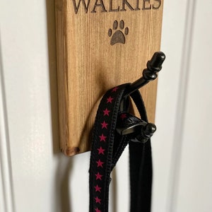 Pu&ograve; includere: Un cartello di legno con la scritta "WALKIES" e un'impronta di zampa, con un gancio nero per appendere un guinzaglio.