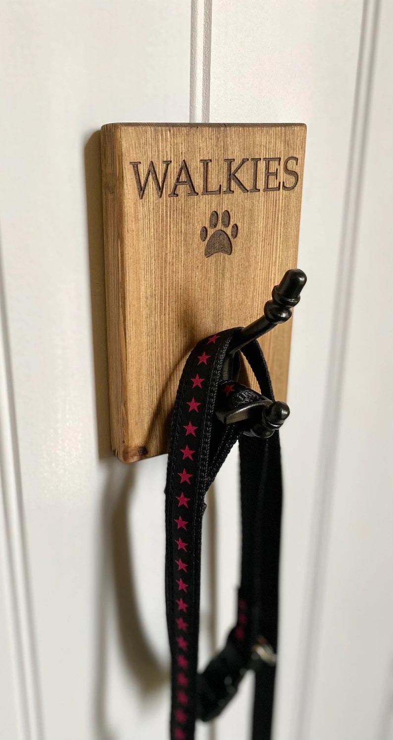 Pu&ograve; includere: Un cartello di legno con la scritta "WALKIES" e un'impronta di zampa, con un gancio nero per appendere un guinzaglio.