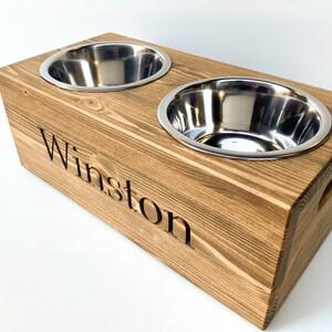 May include: A wooden dog bowl stand with two stainless steel bowls. The stand is stained a dark brown and has the name "Winston" engraved on the front.