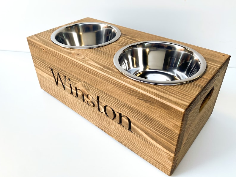 May include: A wooden dog bowl stand with two stainless steel bowls. The stand is stained a dark brown and has the name "Winston" engraved on the front.