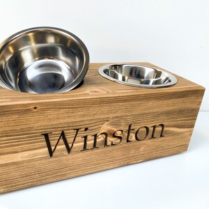 May include: A wooden pet food bowl stand with two stainless steel bowls. The stand is brown and has the name "Winston" engraved on the front.