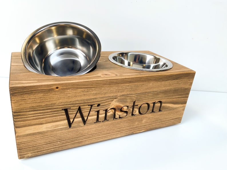 May include: A wooden pet food bowl stand with two stainless steel bowls. The stand is brown and has the name "Winston" engraved on the front.