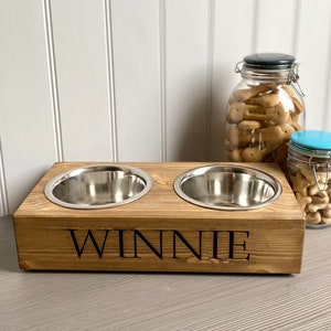 May include: A wooden pet food bowl stand with two stainless steel bowls. The stand is stained brown and has the name "WINNIE" engraved on the front.