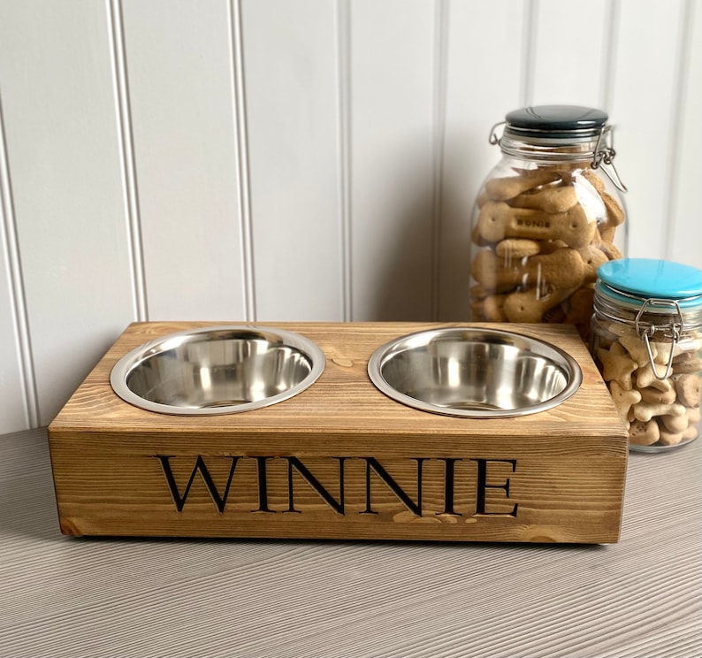 May include: A wooden pet food bowl stand with two stainless steel bowls. The stand is stained brown and has the name "WINNIE" engraved on the front.