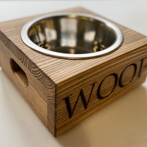 May include: A wooden dog bowl with a stainless steel insert. The bowl is raised on a wooden base and has the word "WOOF" engraved on the side.