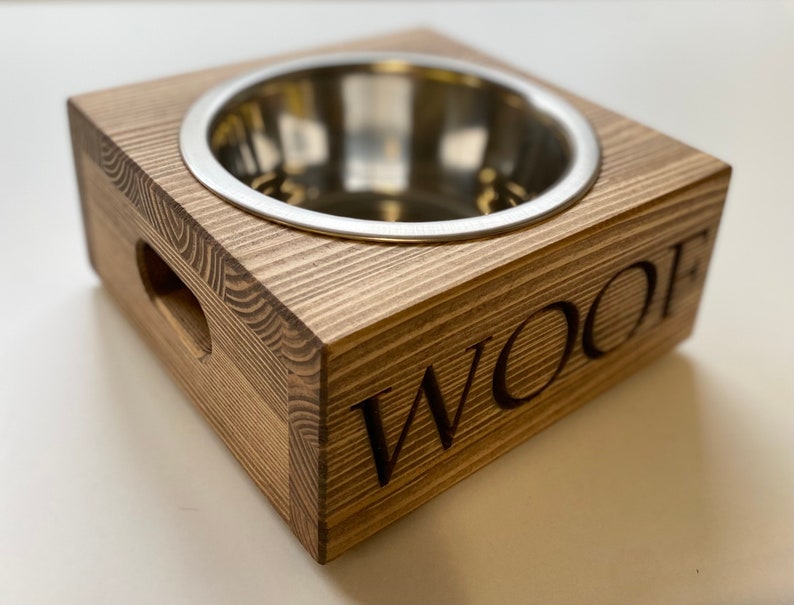 May include: A wooden dog bowl with a stainless steel insert. The bowl is raised on a wooden base and has the word "WOOF" engraved on the side.