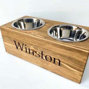 May include: A wooden pet food bowl stand with two stainless steel bowls. The stand is stained a dark brown and has the name "Winston" engraved on the front.