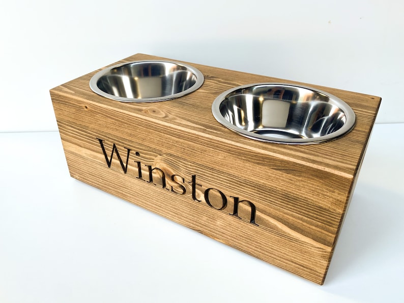May include: A wooden pet food bowl stand with two stainless steel bowls. The stand is stained a dark brown and has the name "Winston" engraved on the front.