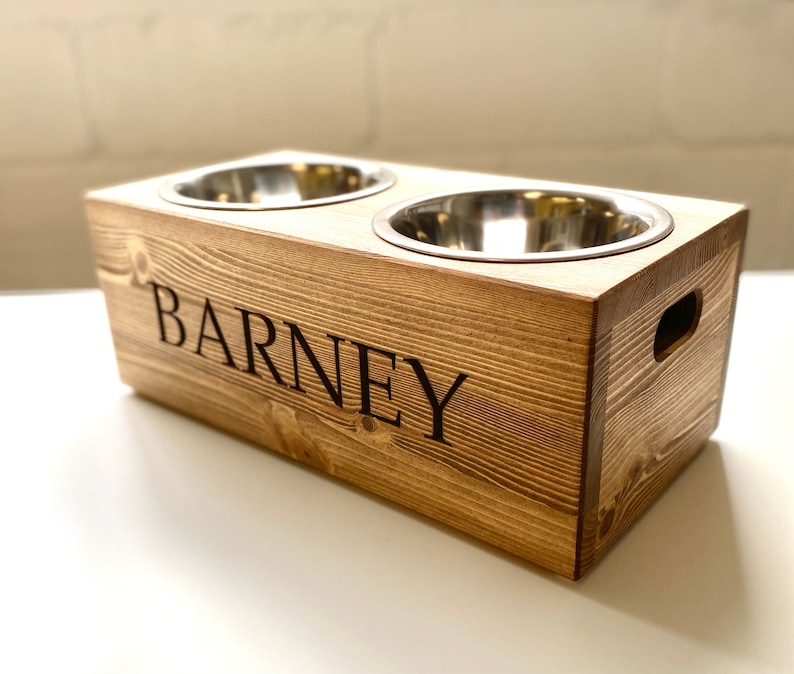May include: A wooden pet food bowl stand with two stainless steel bowls. The stand is stained a dark brown and has the name "BARNEY" engraved on the front.