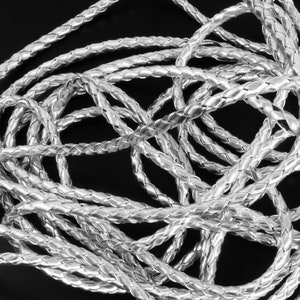 May include: Silver braided cord, a close-up of a silver braided cord, the cord is twisted and coiled, the cord is made of a soft, flexible material, the cord is ideal for jewellery making, crafts, and other projects.