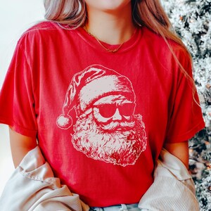 May include: A red t-shirt featuring a white graphic of Santa Claus wearing sunglasses. The Santa illustration includes his hat, beard, and mustache. The shirt is short-sleeved and has a relaxed fit, perfect for the holiday season.