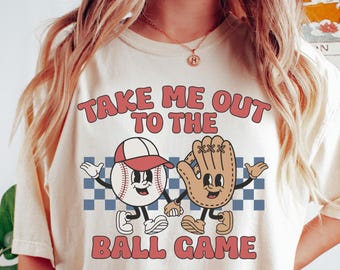 May include: Ivory-colored t-shirt with the text "TAKE ME OUT TO THE BALL GAME" in red and white. The graphic features a baseball and baseball glove with smiling faces, arms, and legs, set against a blue and white checkered background.