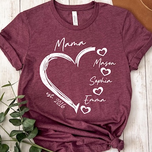 May include: A heather maroon t-shirt featuring a white graphic of a heart outline with the text Mama, est. 2016, and a vertical list of names Mason, Sophia, and Emma, each accompanied by a small white heart icon printed on the front of the garment.