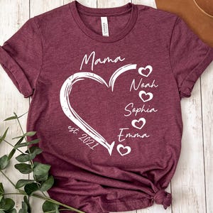 May include: A maroon short-sleeve t-shirt featuring a white graphic of a heart outline with the text Mama, est. 2021, and the names Noah, Sophia, and Emma, each accompanied by a small heart icon, printed on the front of the garment.