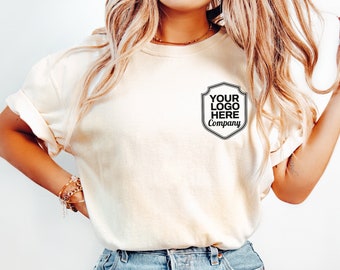 May include: An ivory short-sleeved t-shirt featuring a black graphic on the left chest. The graphic is a shield-shaped emblem containing the text "YOUR LOGO HERE Company" in a bold, sans-serif font. The shirt has a crew neckline and rolled sleeves.