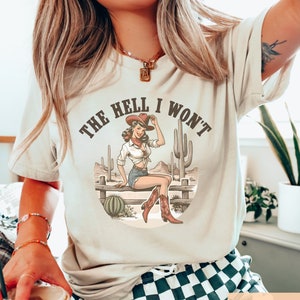 May include: A cream colored t-shirt with a vintage graphic of a woman in a cowboy hat sitting on a fence with the text "The Hell I Won't".