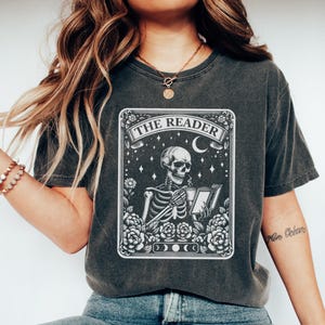 May include: Dark gray t-shirt featuring a detailed graphic of a skeleton reading a book, framed by flowers and the text "THE READER." The design includes stars, a crescent moon, and phases of the moon. The shirt has a vintage, worn look.