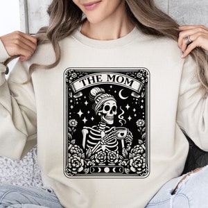 May include: A beige sweatshirt featuring a black and white graphic of a skeleton wearing a beanie and holding a cup of coffee. The graphic is in the style of a tarot card and says "The Mom" at the top.