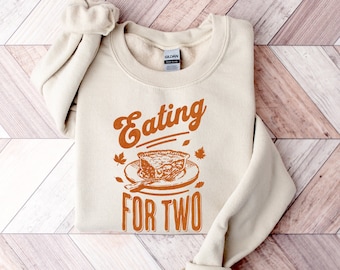 以下が含まれることがあります： オレンジ色の「Eating for Two」という文字が書かれたベージュのトレーナー。文字はビンテージ風のフォントで、プレートにフォークを添えたパイのイラストが添えられています。