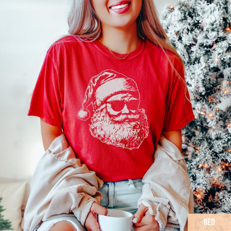 May include: A red t-shirt featuring a white graphic of Santa Claus wearing sunglasses. The Santa illustration includes his hat, beard, and mustache. The shirt is short-sleeved and has a relaxed fit, perfect for the holiday season.