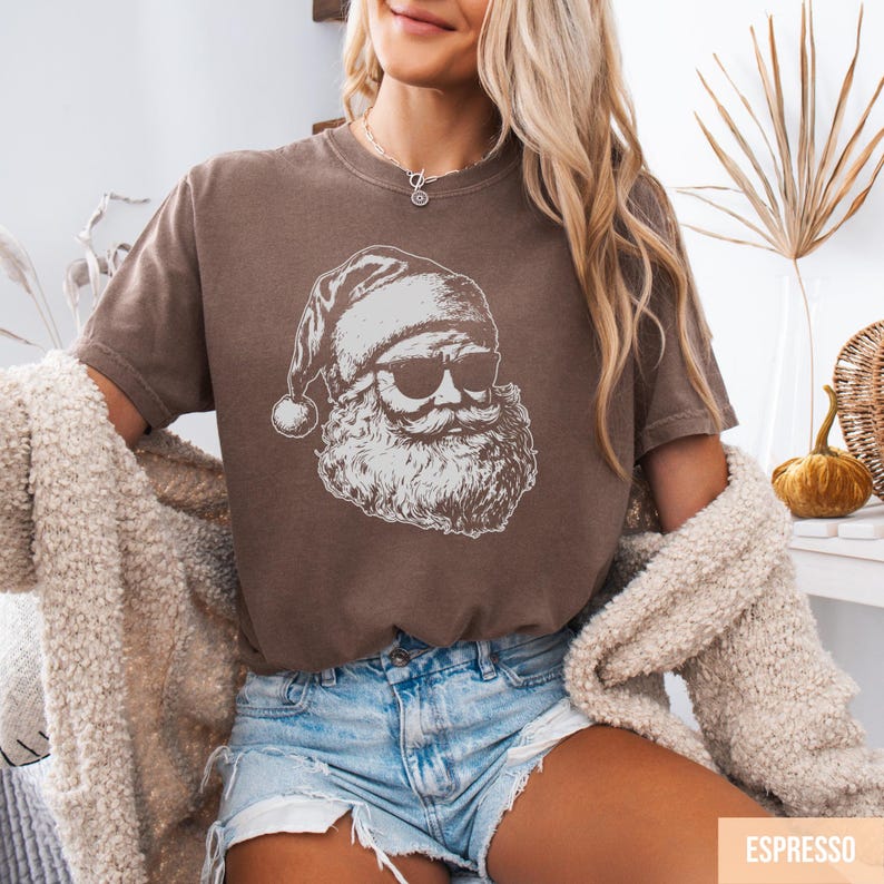 May include: Espresso-colored t-shirt featuring a white graphic of Santa Claus wearing sunglasses. The Santa illustration has a long beard and a traditional hat. The shirt is paired with denim shorts.