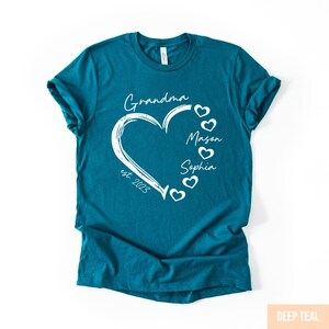 May include: Teal t-shirt with a white heart design. The heart contains the word "Grandma" and the names "Mason" and "Sophia". The text "est. 2023" is also included. The shirt is a solid teal color.